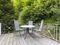 Die Terrasse ist von einem weißen Geländer eingefasst und liegt direkt an einem üppig bewachsenen Wald mit vielen grünen Bäumen und Sträuchern.